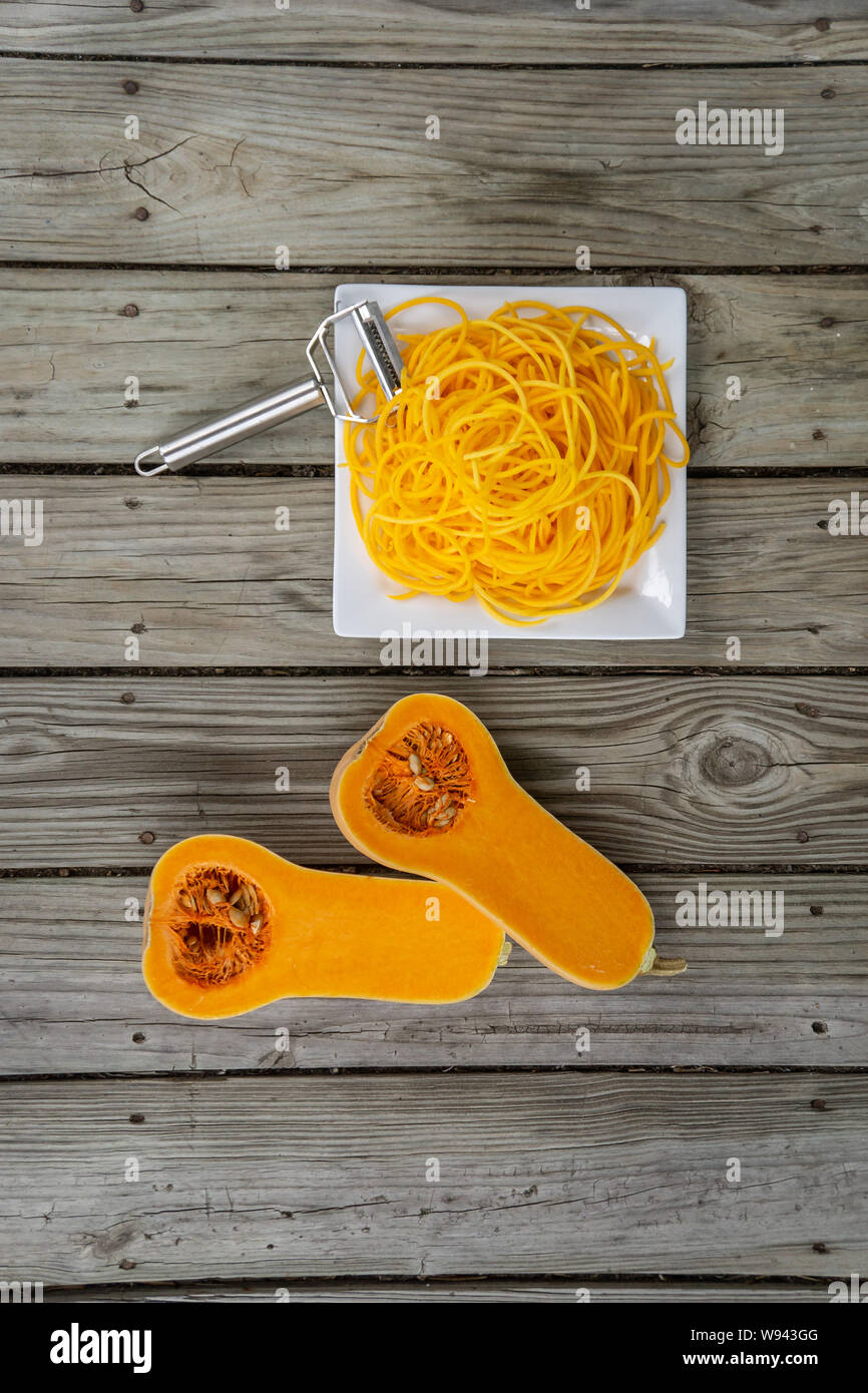 Overhead top view of freshly peeled butternut squash noodles and julienne peeler. Wooden background and white plate for fresh natural vegetable noodle Stock Photo