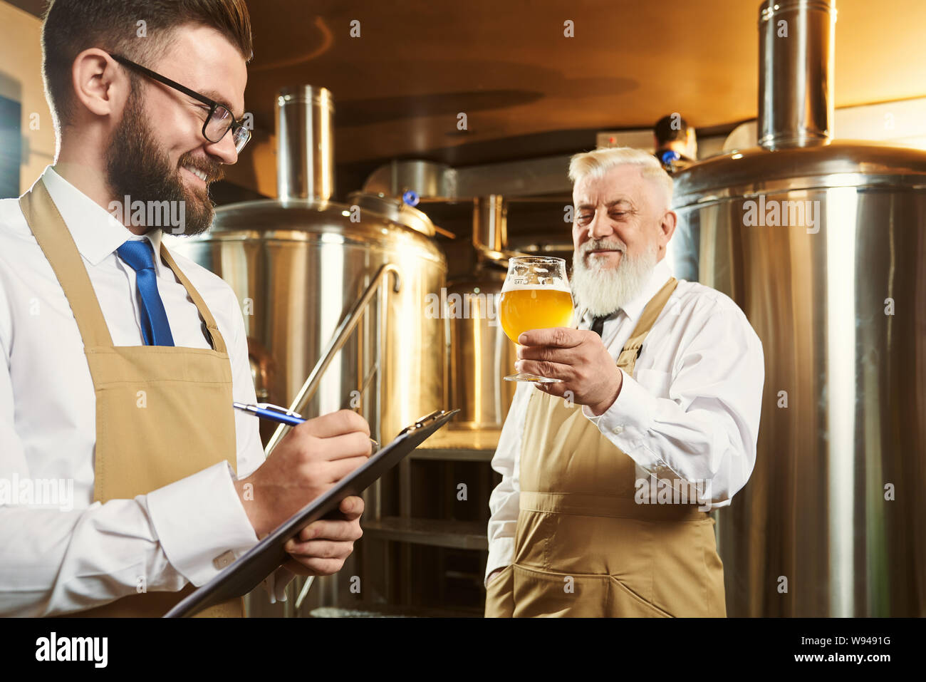Two brewers standing in brewery and examining quality of ale. Bearded man keeping glass of beer while colleague keeping folder and writing down data. Concept of manufacturing. Stock Photo