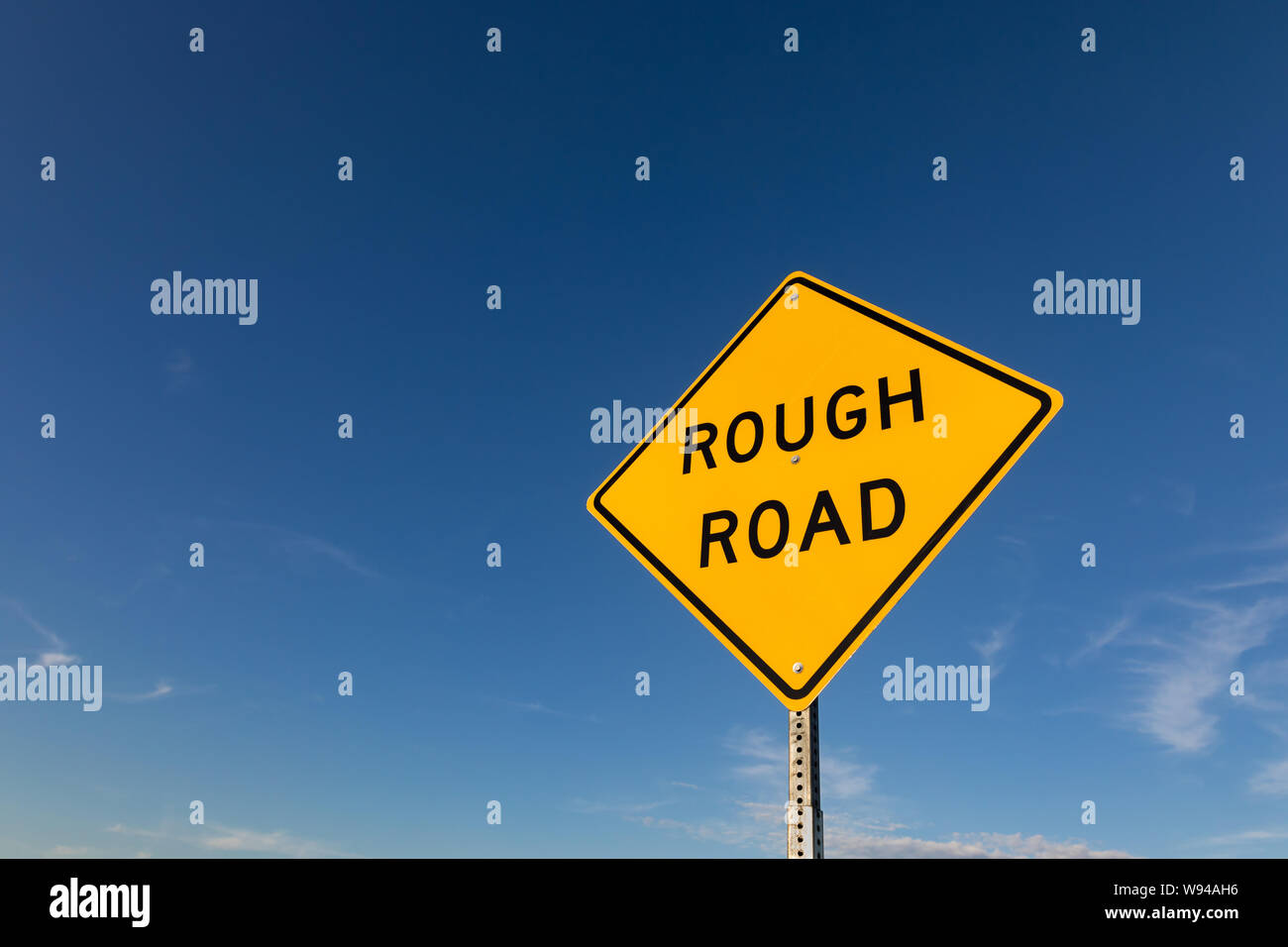 Isolated closeup looking up at yellow rough road warning street sign with blue sky and clouds in background Stock Photo