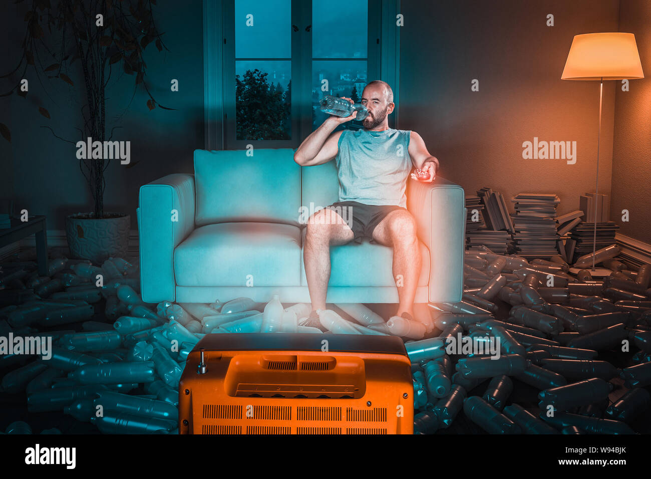 man watches tv on the sofa and drinks from a plastic water bottle, floor full of used bottles. Concept of exaggerated use of plastic and recycling. Stock Photo