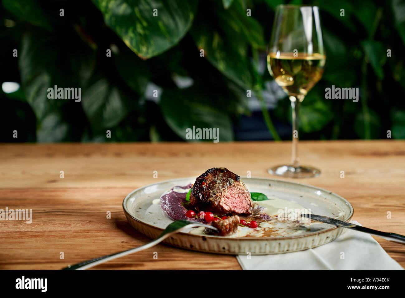 Selective focus of juicy meat served with puree and cranberry on plate in restaurant. Delicious piece of kebab, fork and knife lying near and glass pf champagne staying at background. Concept of meal. Stock Photo
