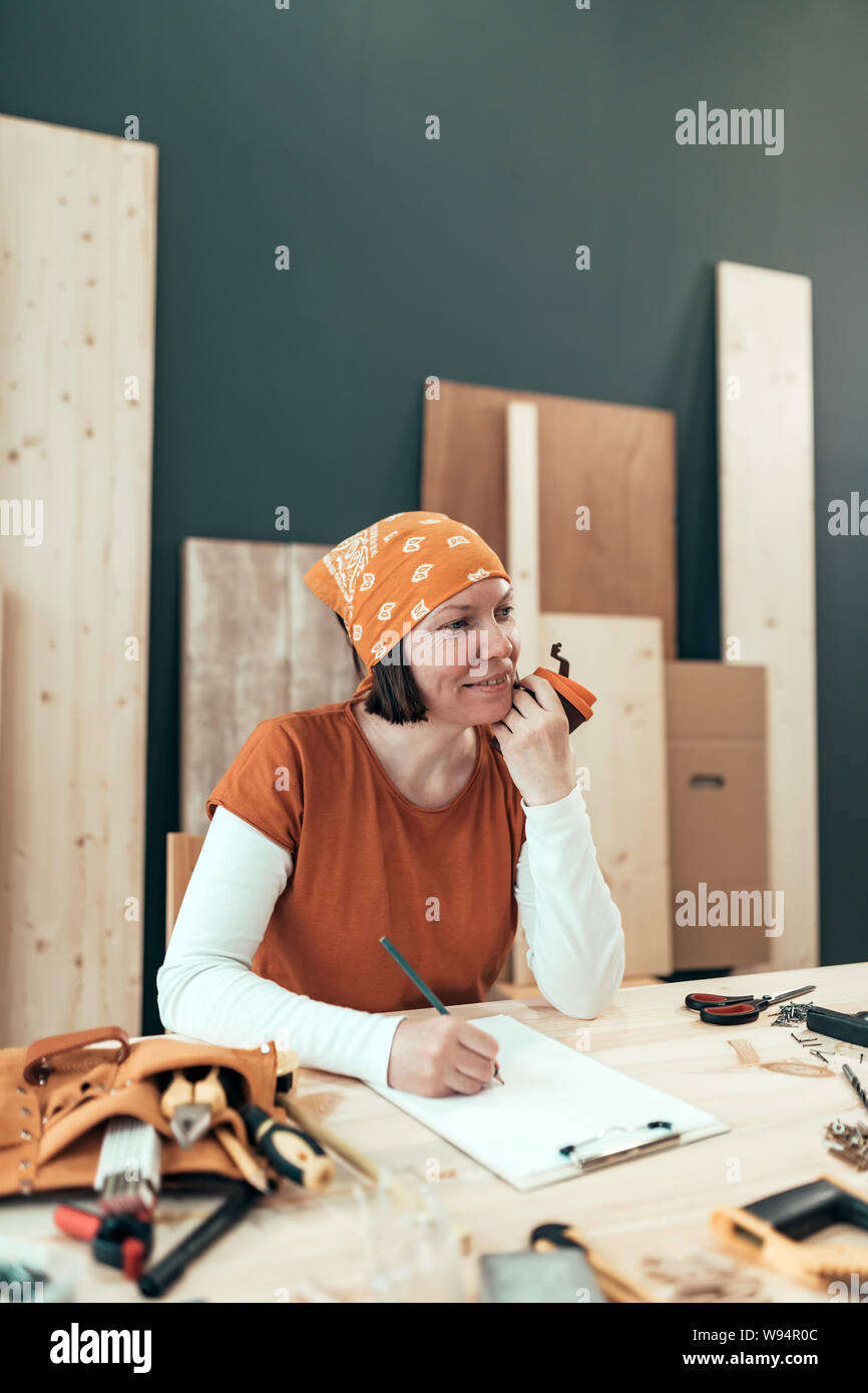 Female carpenter is drinking coffee and planning DIY project in her small business woodwork workshop Stock Photo