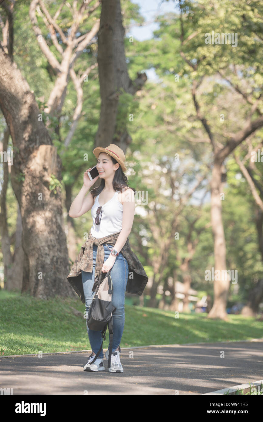 Beautiful asian solo tourist woman  smiling and enjoy taking via mobile phone while walking in public nature park. Vacation travel in summer. Stock Photo