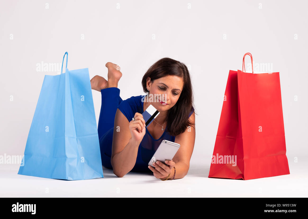 Pretty young Caucasian woman lying on the floor looking at the mobile phone and using the credit card along with a few shopping bags, the concept of o Stock Photo