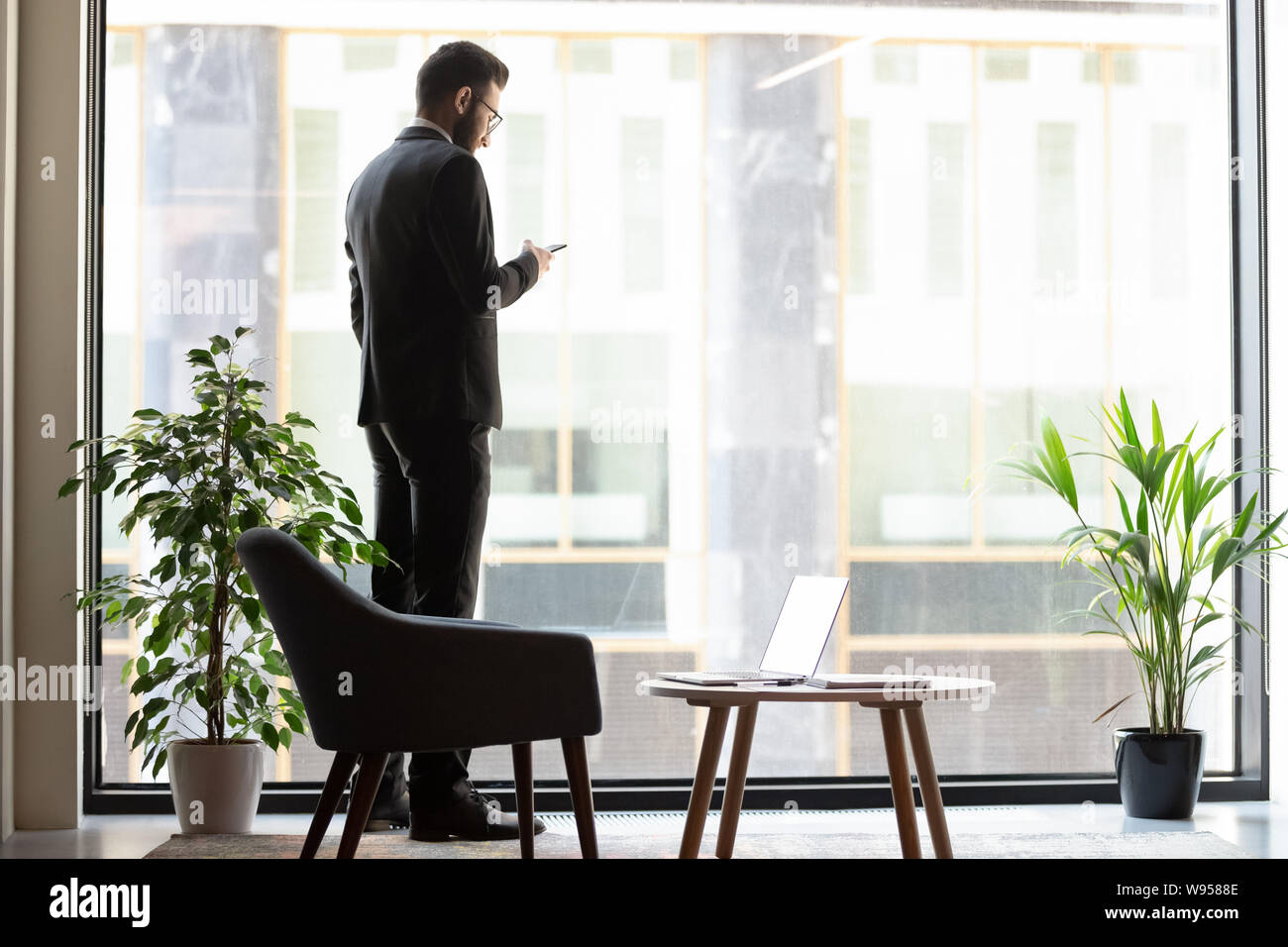 Confident businessman standing near window, using phone Stock Photo