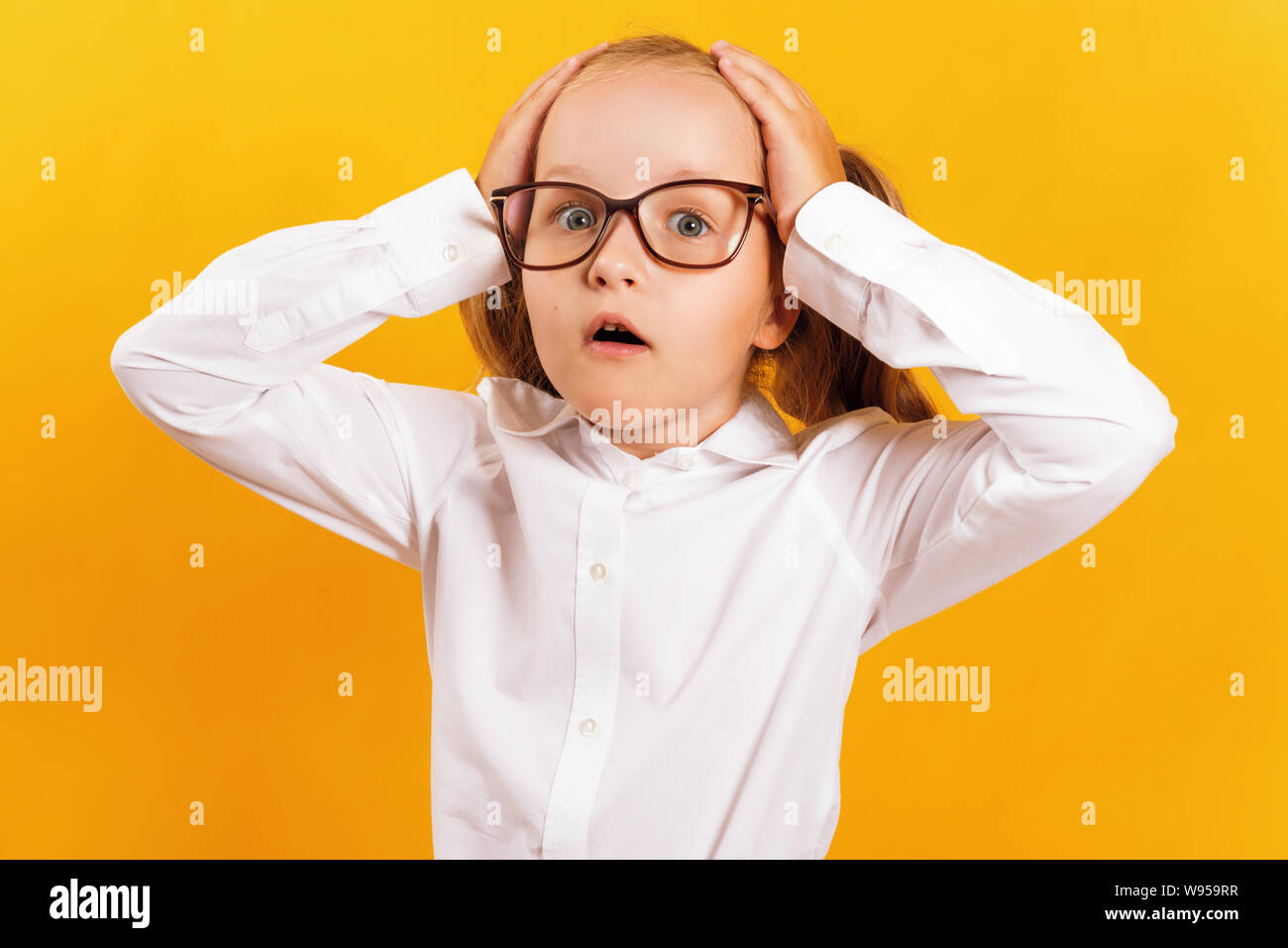 Portrait of a cheerful little girl in glasses on a yellow background. The child is shocked and holds his hands behind his head. Stock Photo