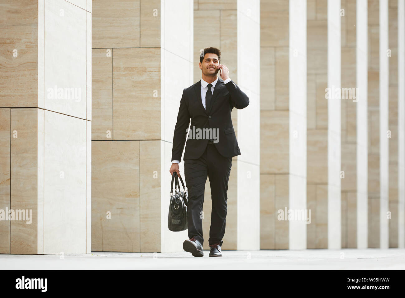 Young businessman in black suit carrying bag and has a business conversation on mobile phone while walking in the city Stock Photo