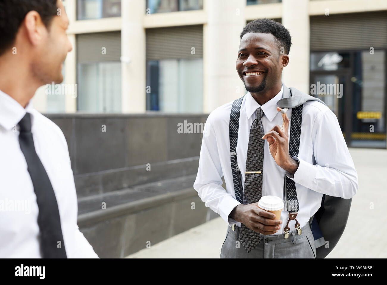 Happy African businessman holding cup of coffee talking to his colleague and smiling while standing outdoors Stock Photo