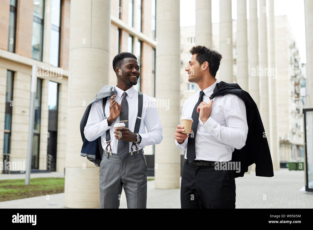 Two multiethnic office workers smiling and talking to each other while walking along the street and drinking coffee from disposable cups Stock Photo
