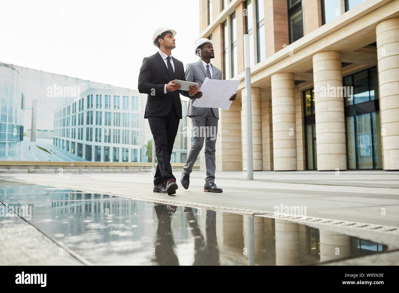 Two multiethnic architects in formal clothing and hardhats holding blueprint walking along the street and looking at modern building Stock Photo