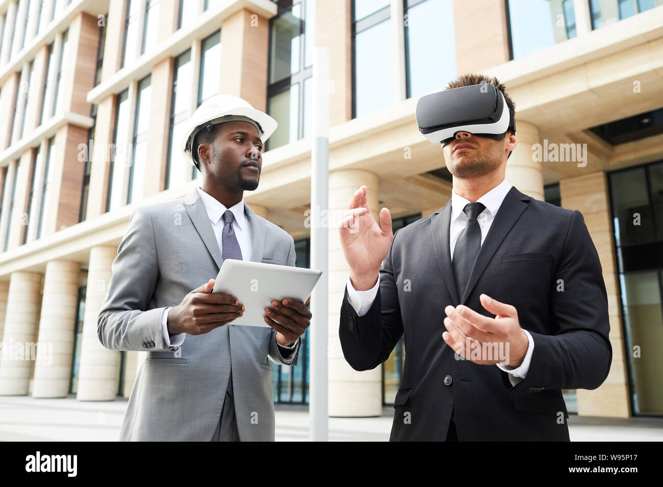 Young businessman wearing virtual reality glasses standing outdoors together with his colleague who using digital tablet in the city Stock Photo