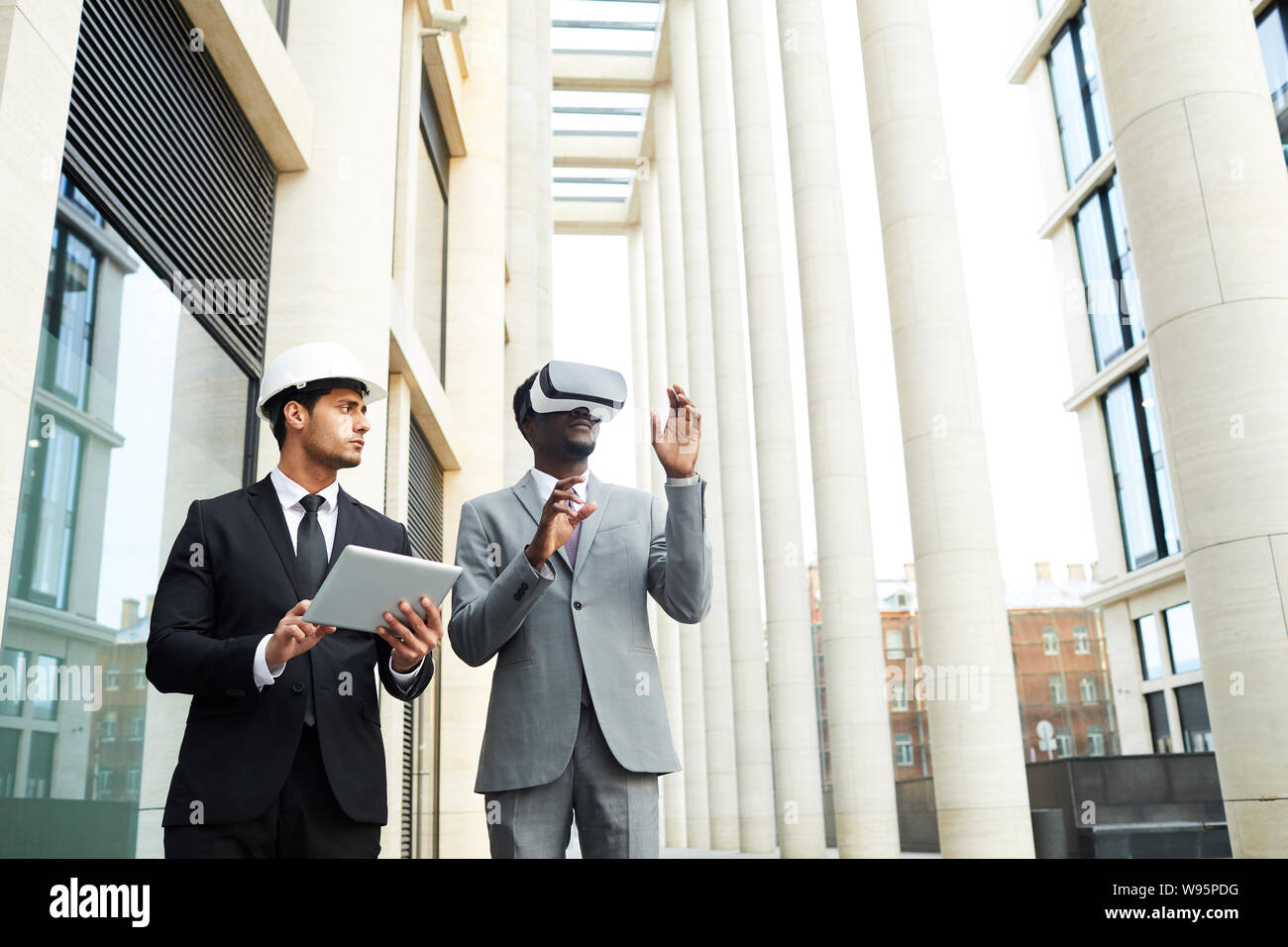 Young businessman in hardhat and suit using tablet pc and looking at his colleague while he wearing virtual reality goggles and walking in the city Stock Photo