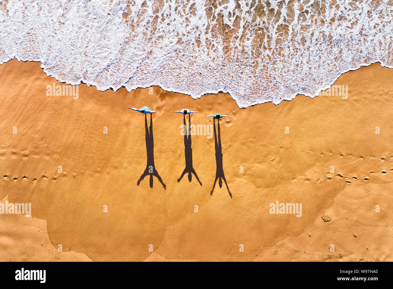 Three people facing rolling waves of Pacific ocean on a clean wide sandy beach during morning exercise in aerial top down view. Stock Photo