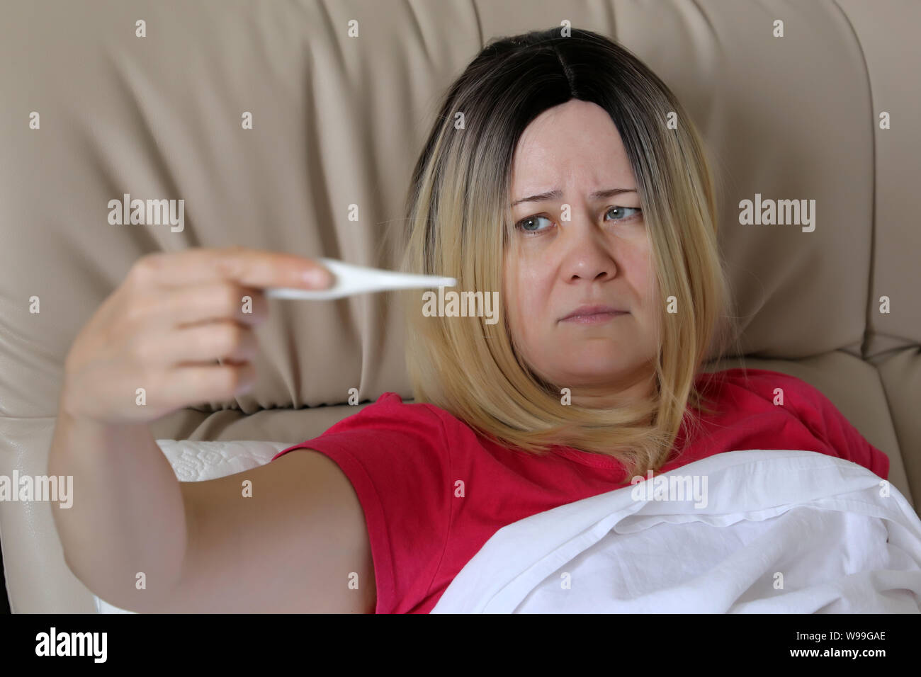 Sick woman measuring body temperature lying in a bed, digital thermometer in female hand. Concept of fever, cold and flu, illness, head pain Stock Photo