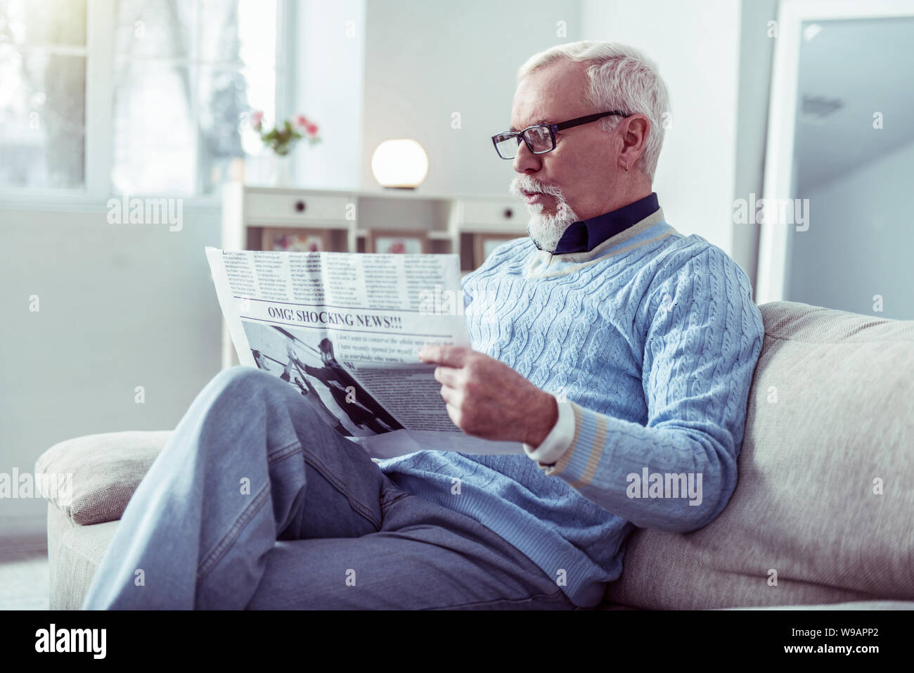 Bearded retired man wearing glasses sitting on sofa and reading newspaper Stock Photo