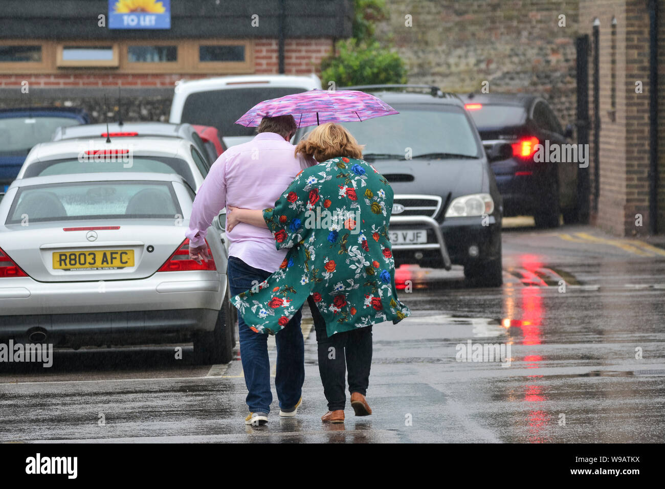 A couple of people caught out in the rain sharing an umbrella, heading for a car in a car park on a wet day in the UK. Raining in UK. Stock Photo