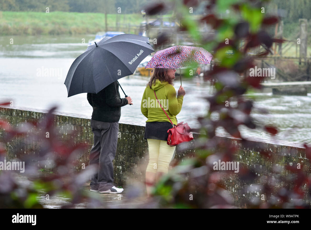 A couple standing in the rain by a river holding umbrellas on a wet day in the UK. Raining in Autumn in the UK. Stock Photo