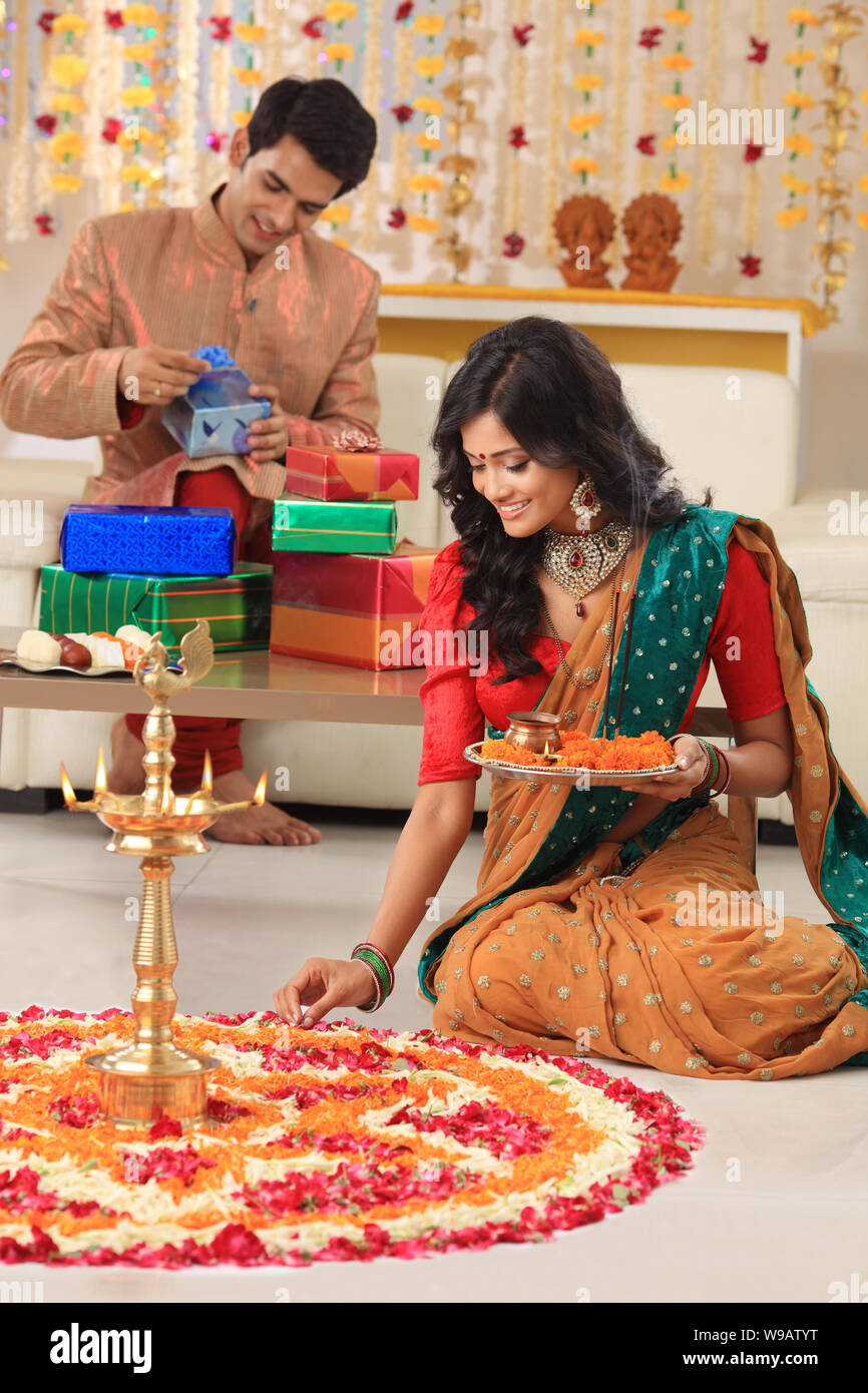 Young woman making rangoli with her husband packing gifts in the background Stock Photo