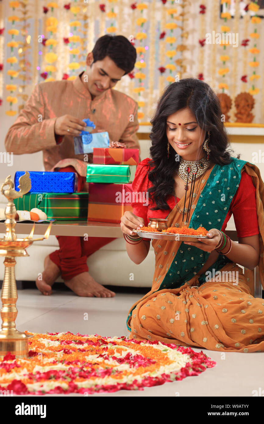 Young woman making rangoli with her husband packing gifts in the background Stock Photo