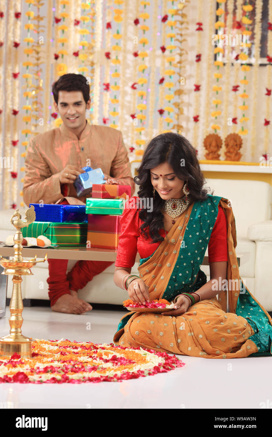Young woman making rangoli with her husband packing gifts in the background Stock Photo