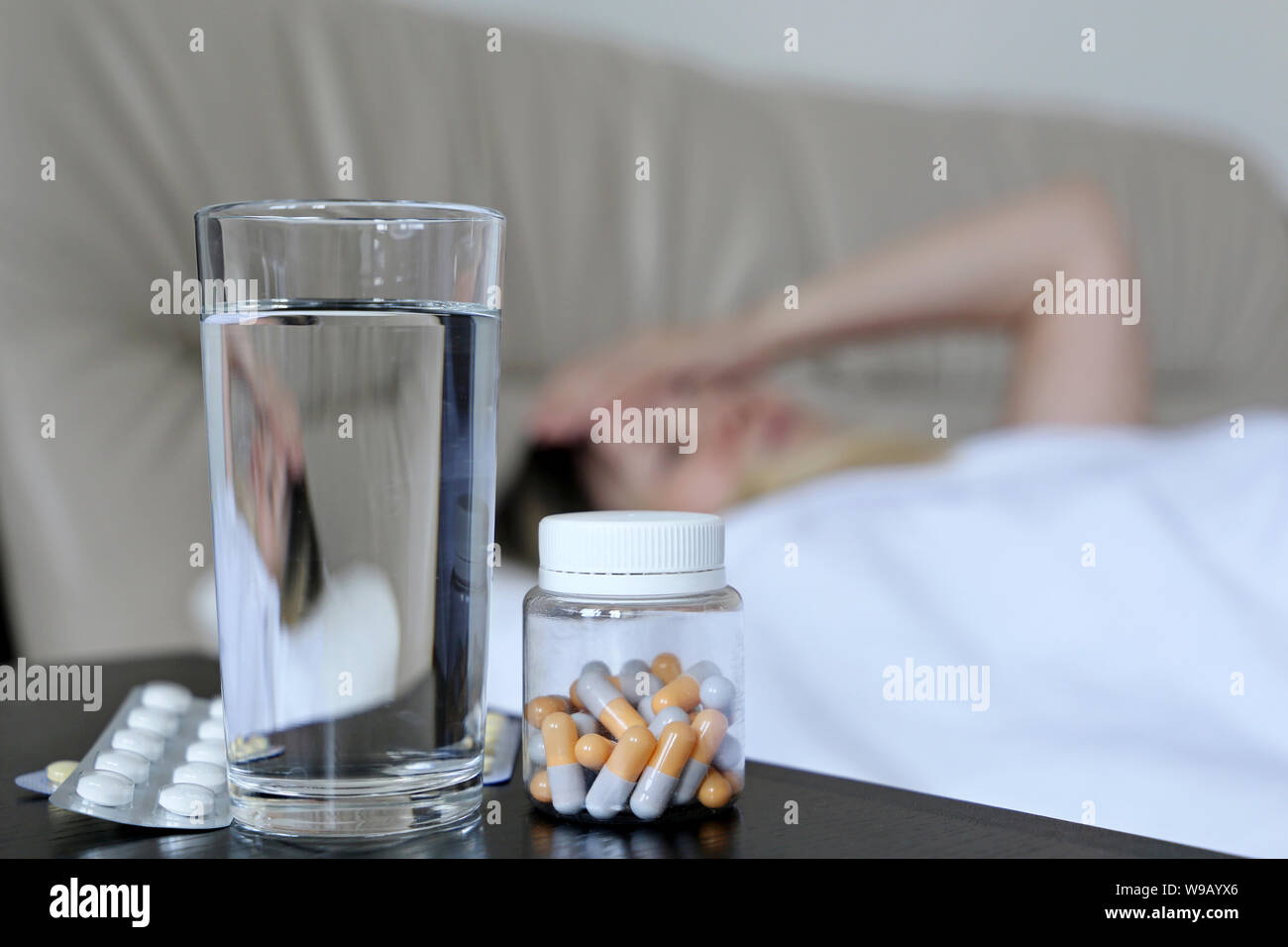 Cold and flu season, pills and water glass on background of sick woman lying in a bed. Concept of illness, fever, hot temperature Stock Photo