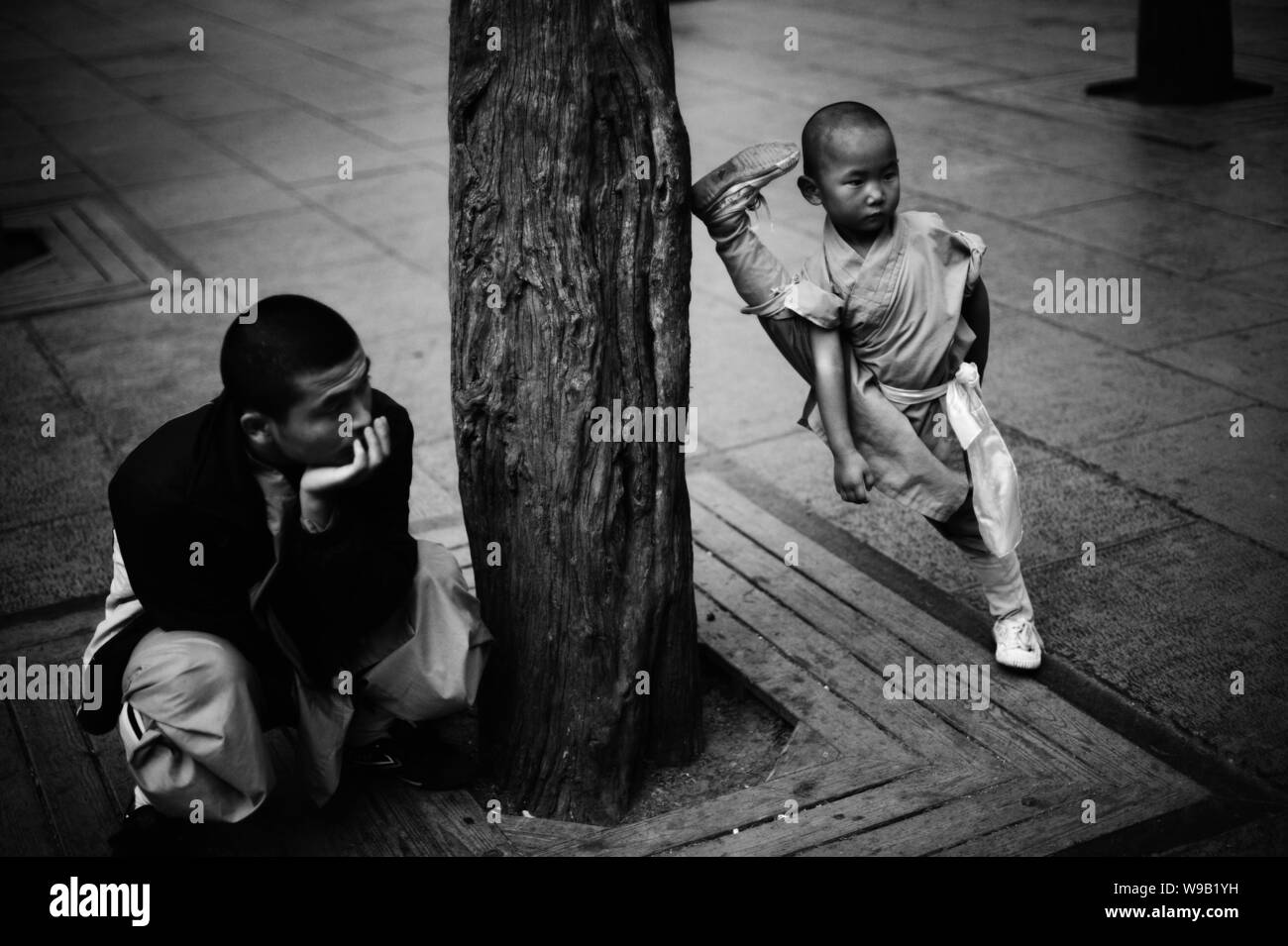 A little boy from the Shaolin Kungfu Training Base opens his legs wide against a tree next to his master while practising kungfu at the Shaolin Temple Stock Photo