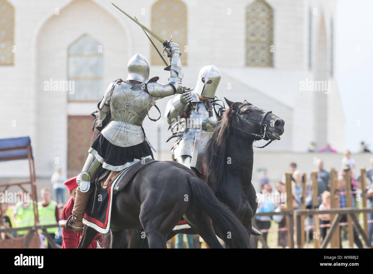 Two men knights riding horses on the battlefield and having a fight - people watching behind the fence Stock Photo