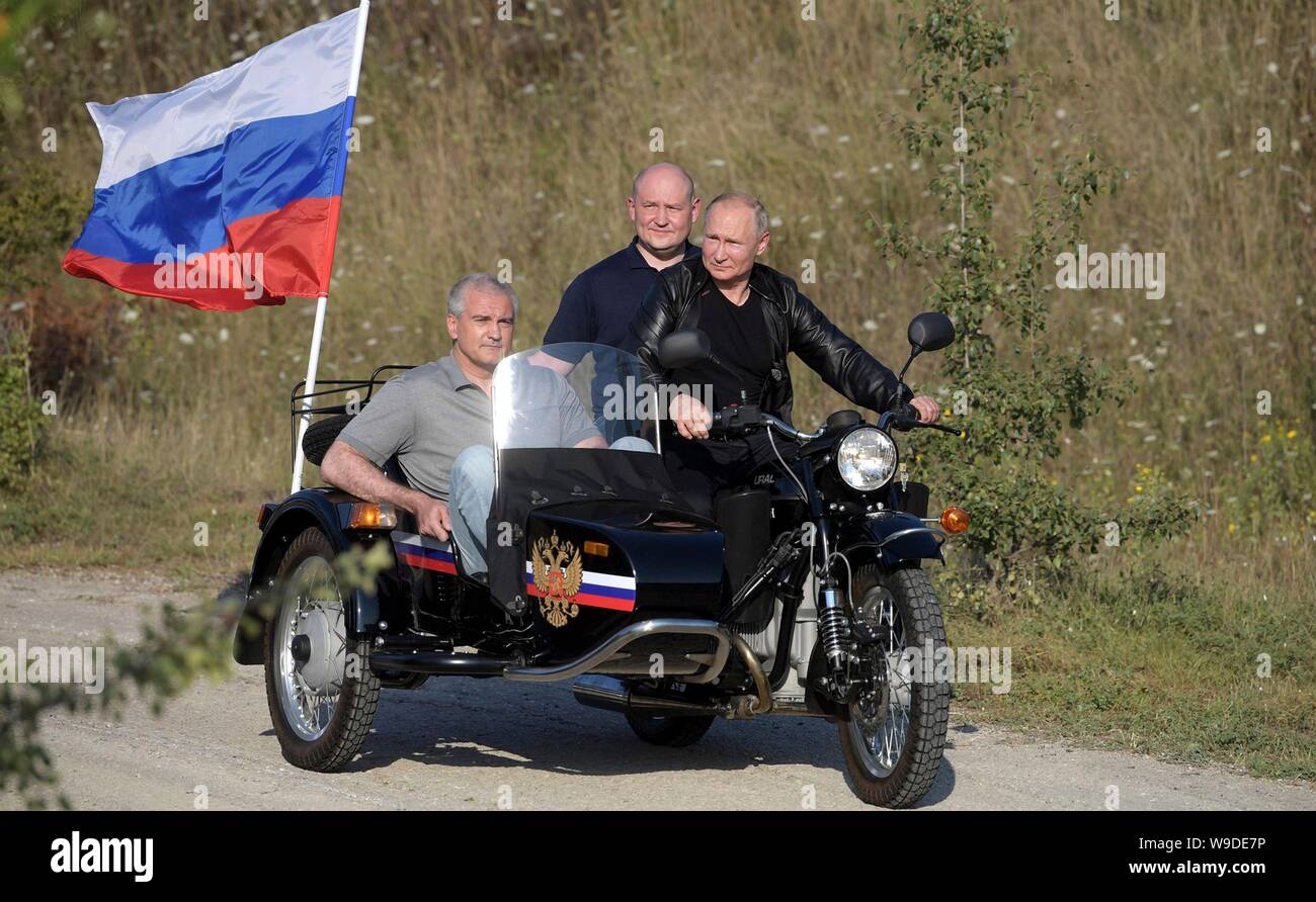 Russian President Vladimir Putin, right, rides a motorcycle and sidecar with the Night Wolves biker gang on their way to the Babylon Shadow bike show and camp August 10, 2019 near Sevastopol, Crimea, Russia. Riding behind Putin in pillion position on the motorcycle is Acting Governor of Sevastopol Mikhail Razvozhayev and Head of the Republic of Crimea Sergei Aksyonov is in the sidecar. Stock Photo