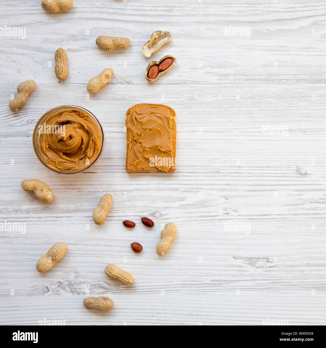 Toast, bowl of peanut butter and peanuts in shells on a white wooden table, overhead view. From above, flat lay, top view. Space for text. Stock Photo