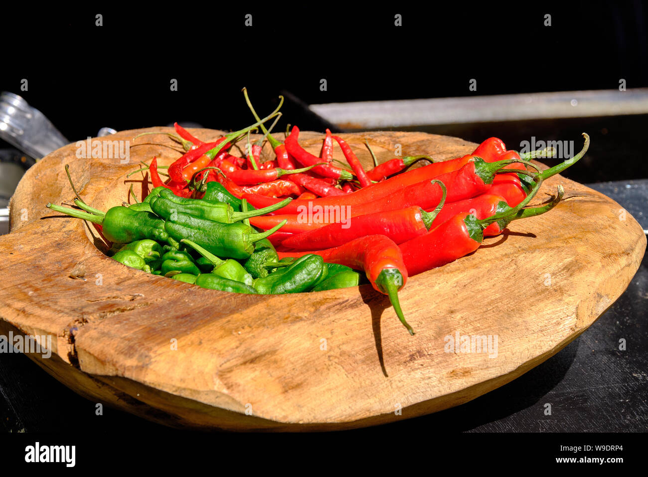 Wooden bowl on black counter filled with green and red chilies on outside counter in bright sunny light Stock Photo
