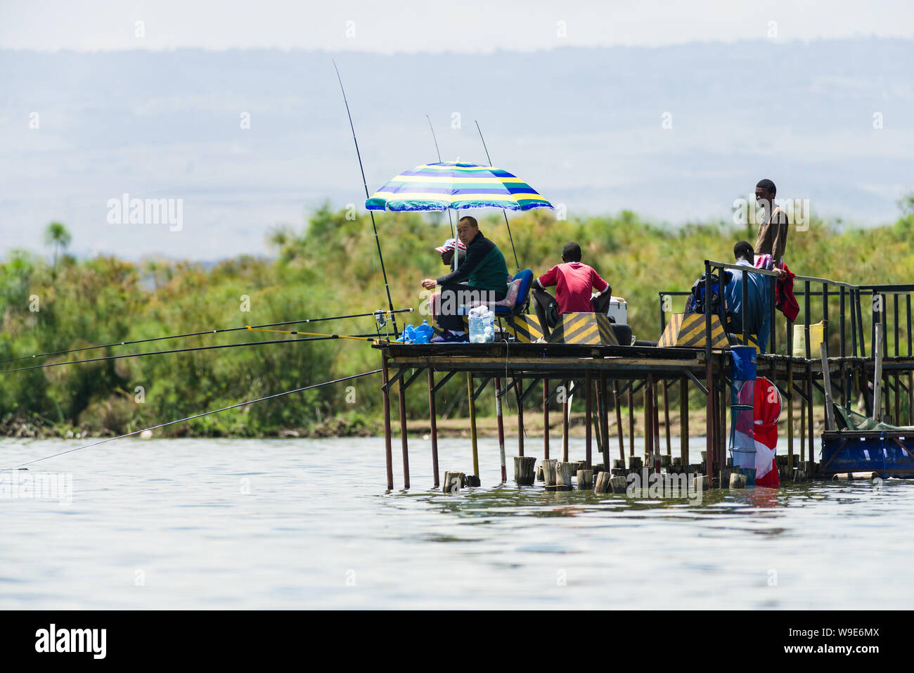 A Chinese man sits fishing with local Kenyan men on a jetty , lake Naivasha, Kenya, East Africa Stock Photo