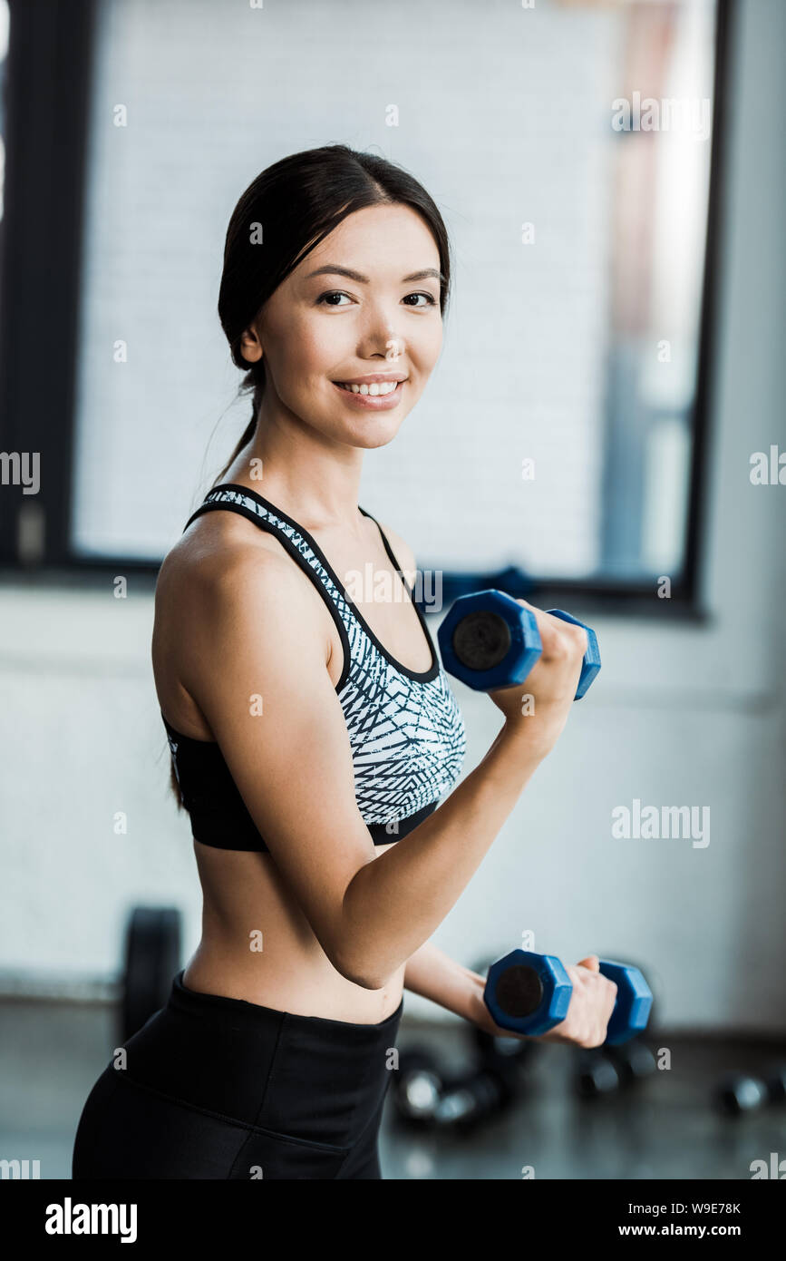 positive and sporty woman holding dumbbells while working out in gym