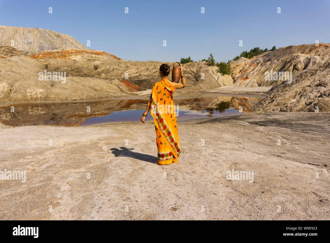 a girl in a sari with a pitcher on her shoulder goes for water to a dirty lake in a deserted landscape Stock Photo