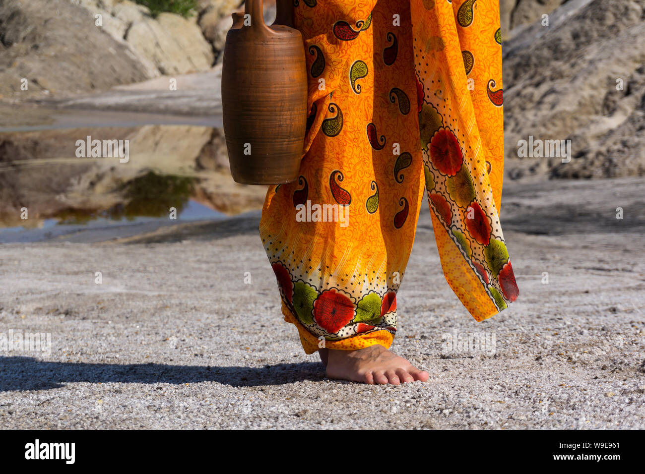 a woman in ethnic dress walking for water with ceramic jug in desert landscape Stock Photo