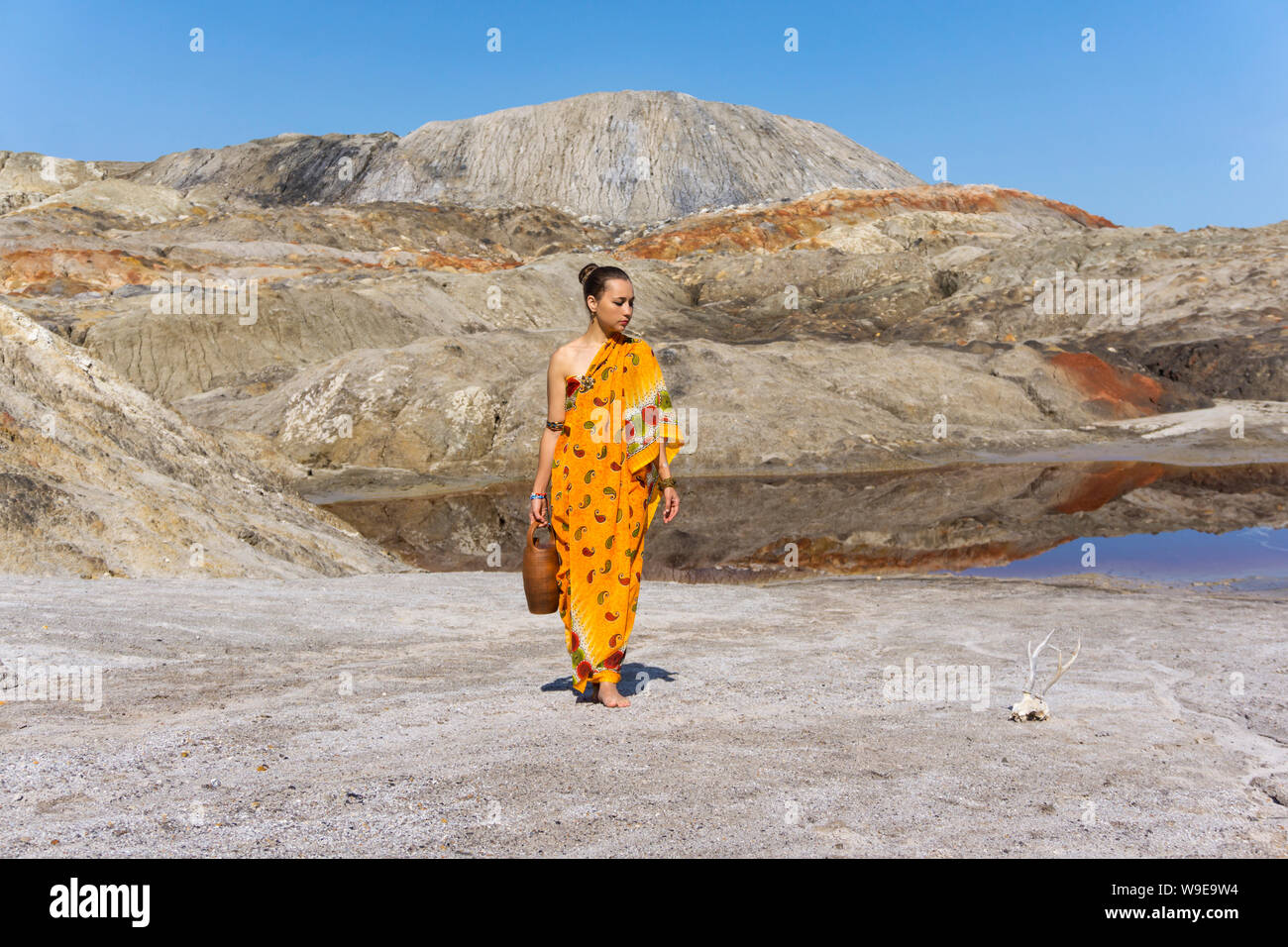 young woman in a sari with a jug comes from a pond in an arid, lifeless area, looking thoughtfully at the skull of an animal Stock Photo