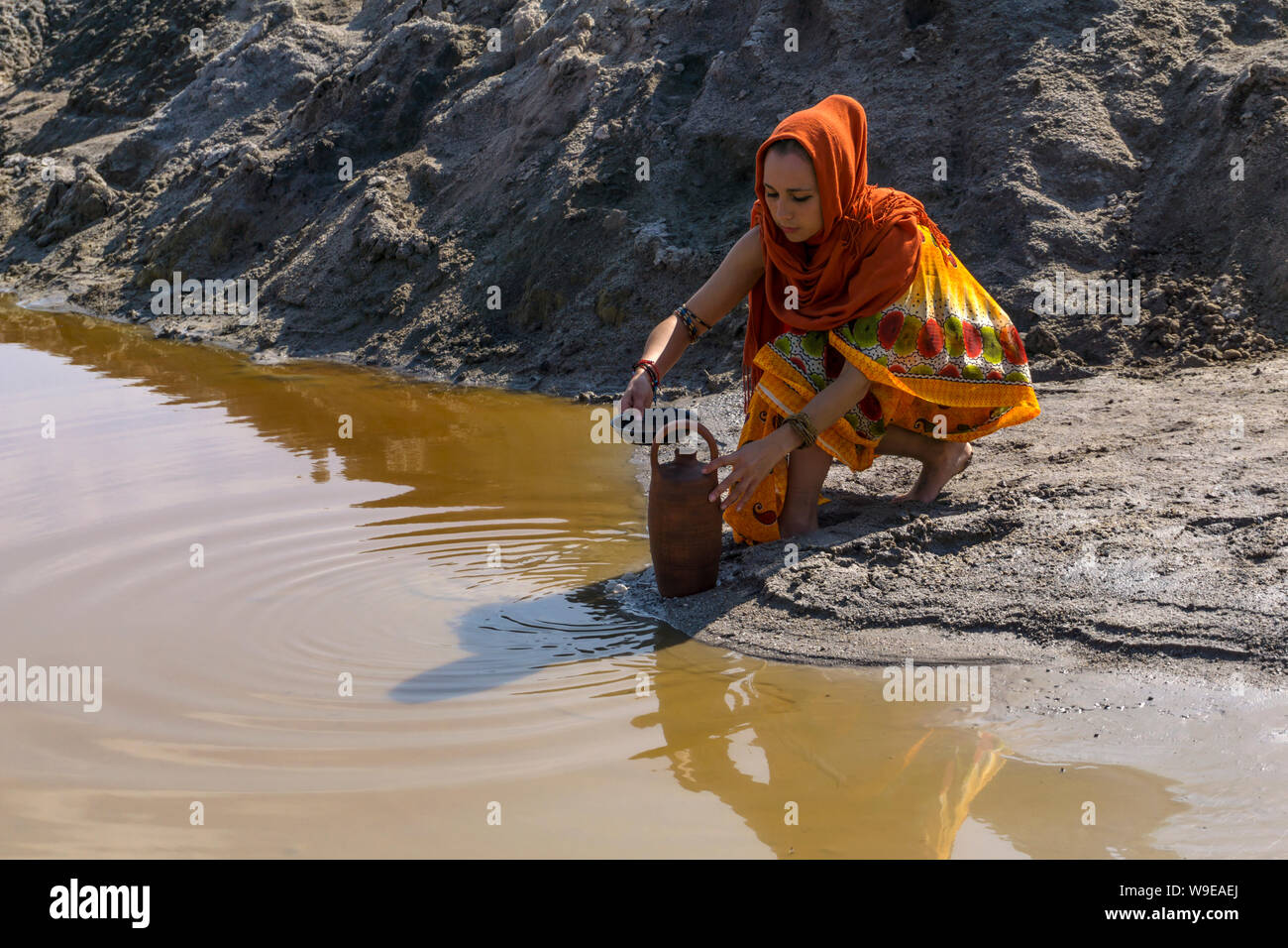 girl of oriental appearance in sari and hijab fills the pitcher with water from a dirty source in the desert Stock Photo