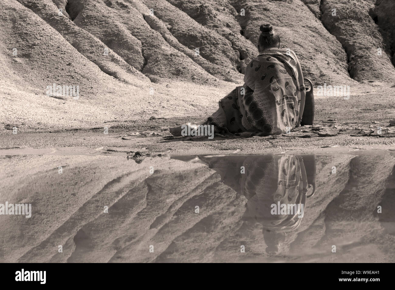 young woman in ethnic attire sits with a jug on the shore of a lake in a deserted valley Stock Photo