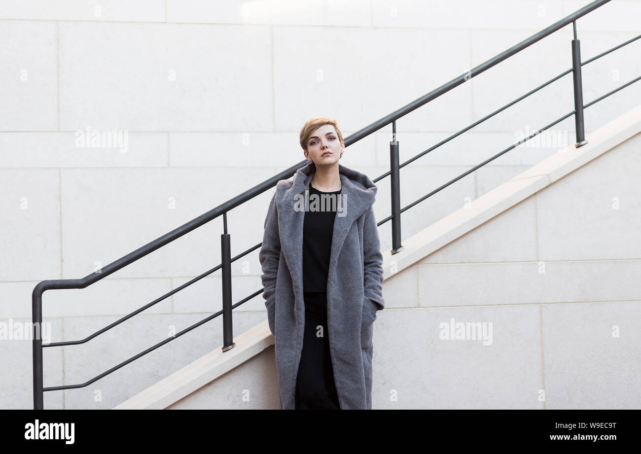 woman with short tousled hair in gray coat on stairs background Stock Photo