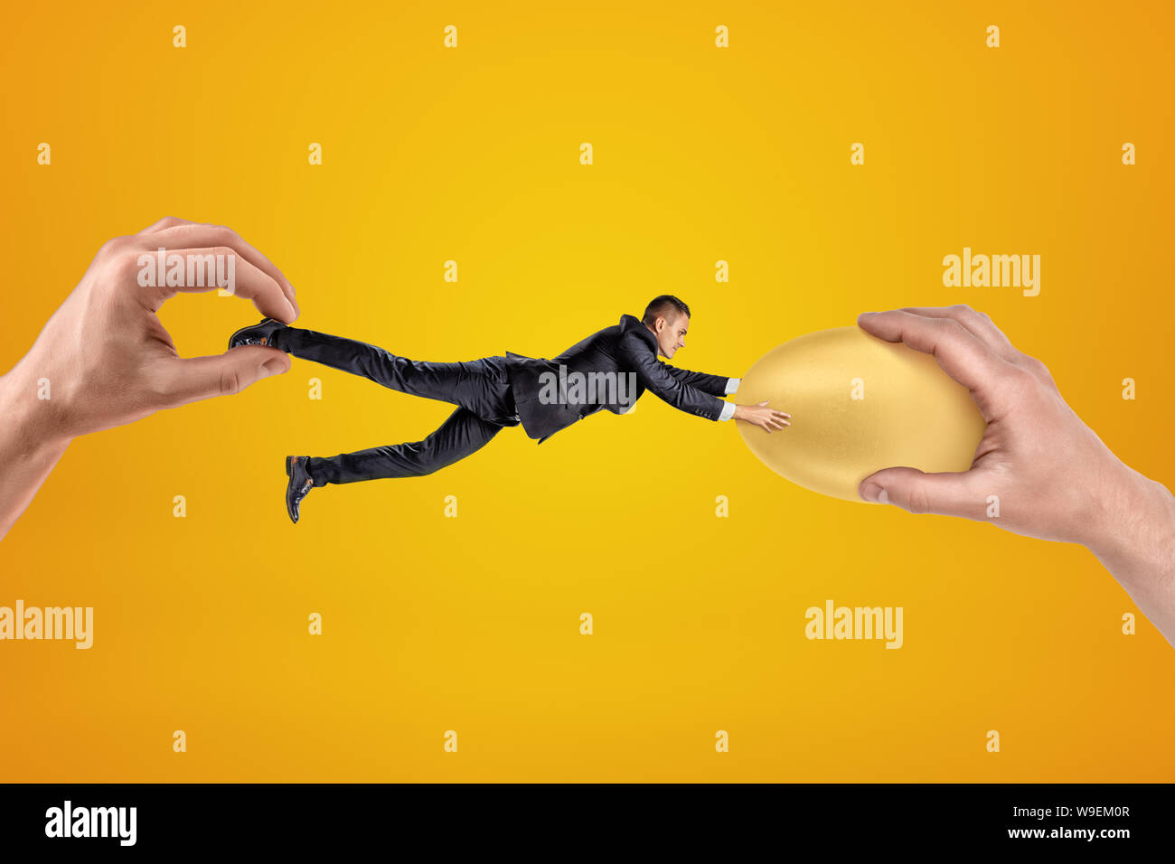 Small businessman being pulled in opposite directions by two big hands, and clutching with his both hands at gold egg in one of the hands. Stock Photo