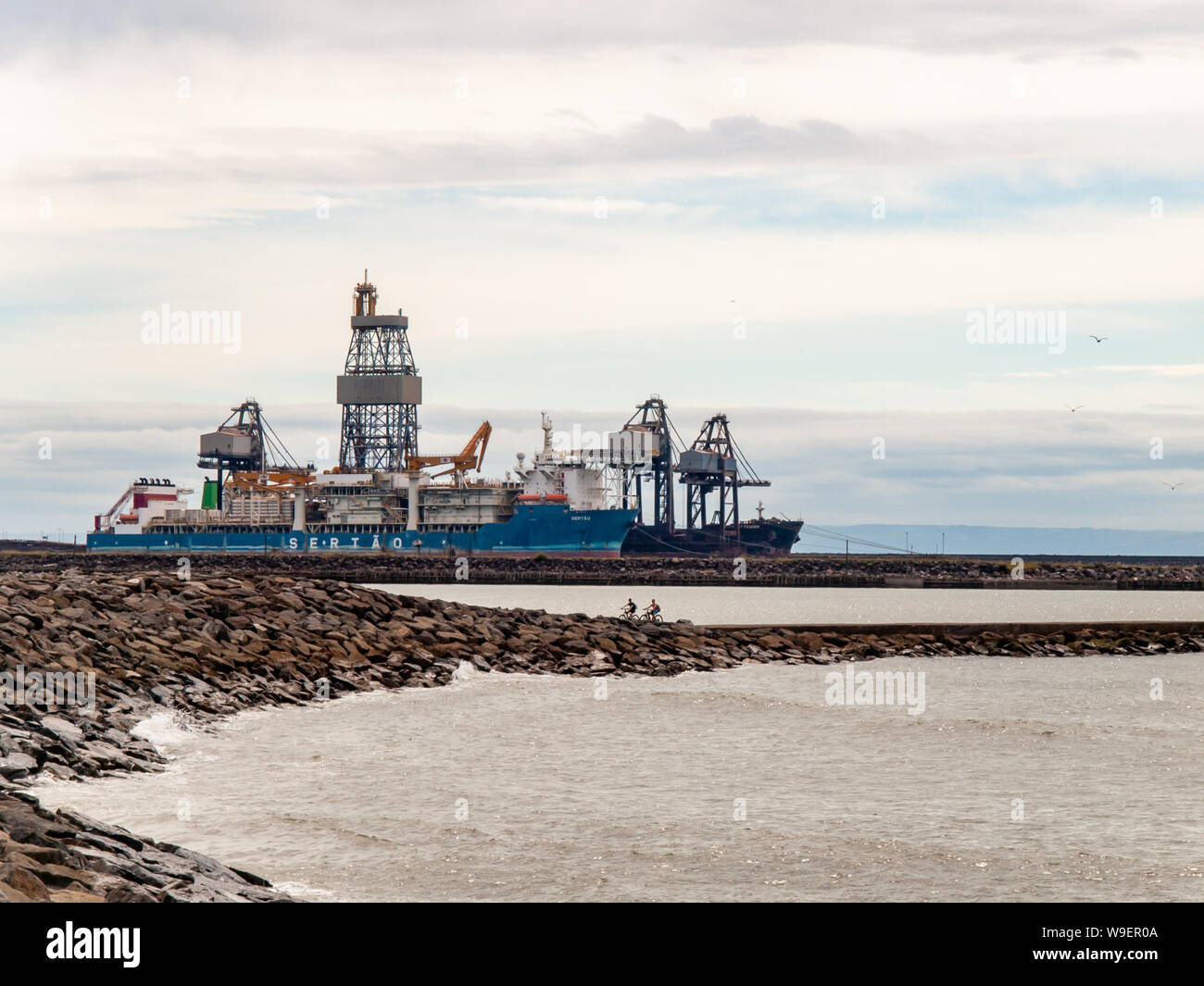 The deep water drilling vessel Sertao at dock in Port Talbot in South Wales. Two mountain bike riders can be seen on the breakwater. Wales, UK Stock Photo