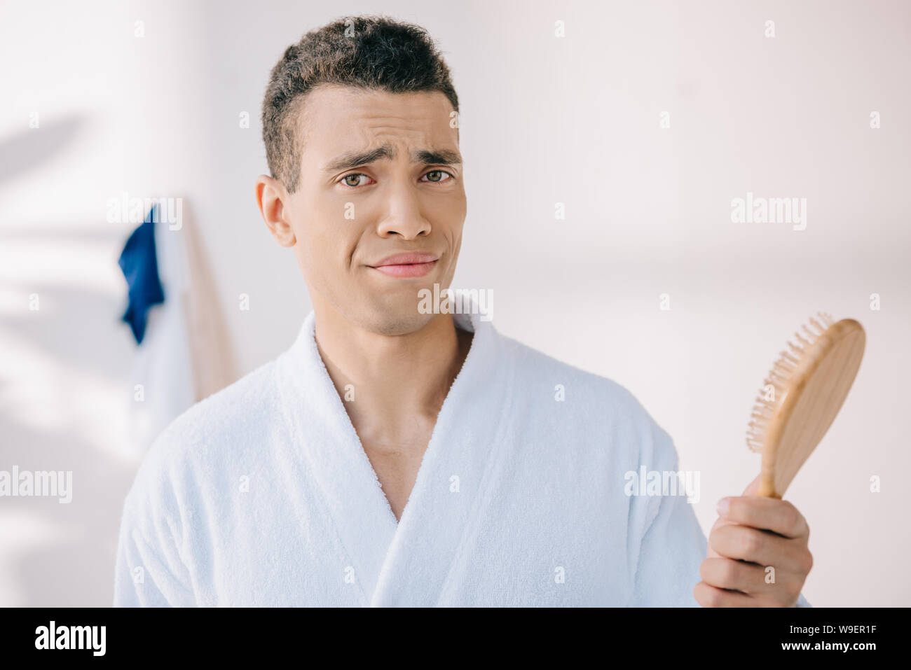 disappointed young man in bathrobe holding hairbrush, looking at camera and making grimace Stock Photo