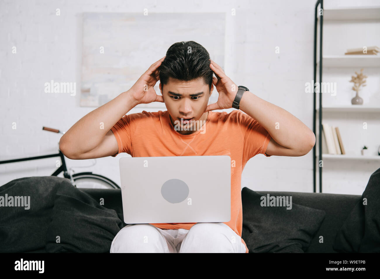 shocked asian man holding hands near head while sitting on sofa and using laptop Stock Photo