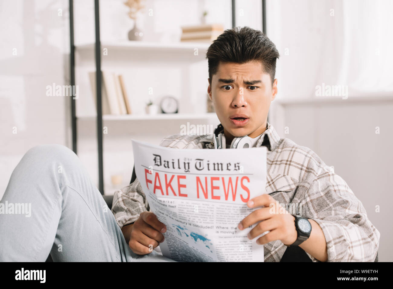 shocked asian man reading fake news newspaper while sitting on couch at ...