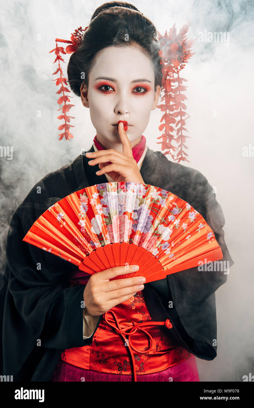 beautiful geisha in black kimono with flowers in hair holding hand fan and showing shh gesture in smoke Stock Photo