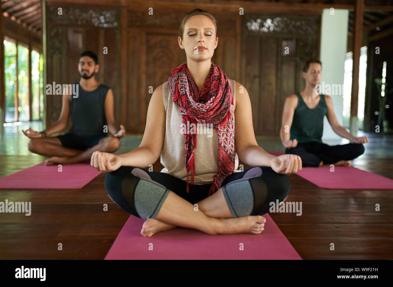Beautiful woman teaching meditation to two multi-ethnic men in lotus pose on yoga mats in traditional temple in Bali Indonesia Stock Photo