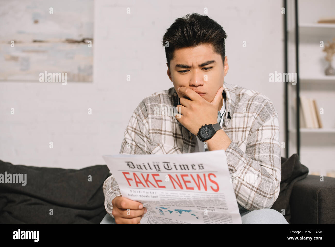 serious asian man reading fake news newspaper while sitting on sofa at ...