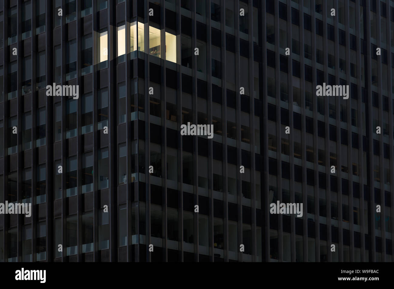 Corner office with lights on and windows glowing in a darkened office building at night indicating someone working overtime late at night Stock Photo