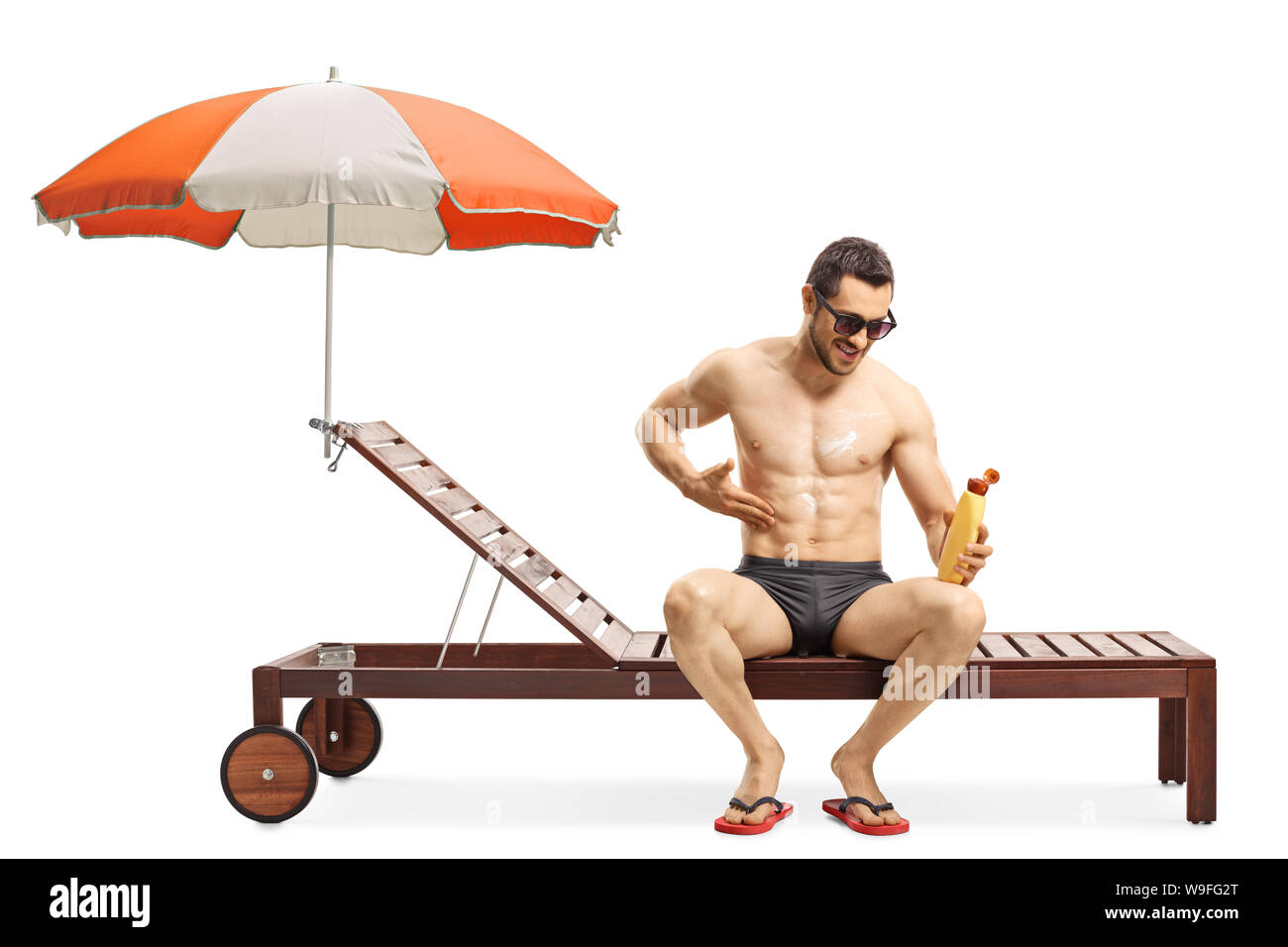 Full length shot of a handsome fit man applying sun cream and sitting on a sunbed under umbrella isolated on white background Stock Photo