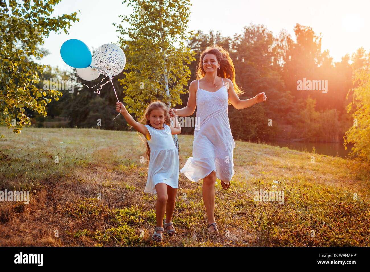 Happy little girl running with mother and holding baloons in hand. Family having fun in summer park at sunset. International Childrens Day Stock Photo