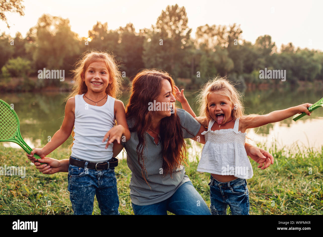 Family having fun after playing badminton by summer river at sunset. Mother laughing and grimacing with daughters outdoors. International Childrens Da Stock Photo